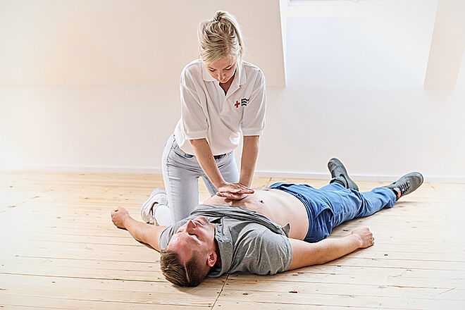 Auf dem Boden liegt ein Mann auf dem Rücken, die Arme seitlich ausgebreitet; darüber beugt sich eine Frau, die eine Herz-Druck-Massage demonstriert
