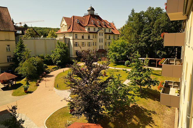 Im Vordergrund sieht man Büsche und Rasenflächen des kleinen Parks, im Hintergrund sieht man Haus Brückenbogen mit dem Kuppeltürmchen